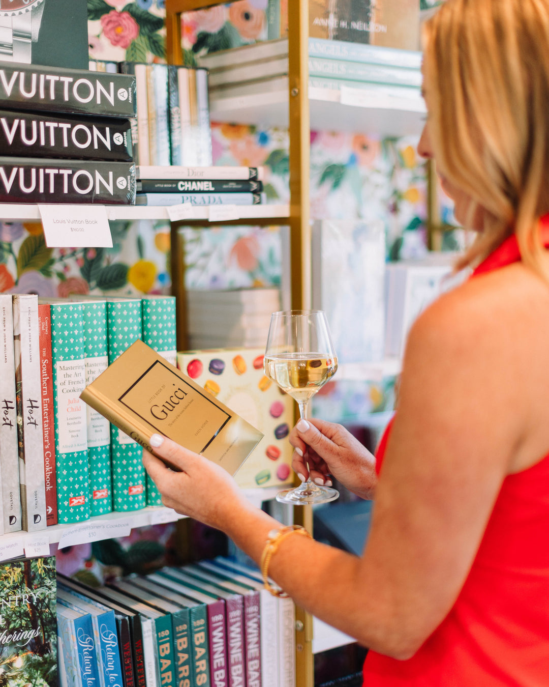 Woman in a red dress holding a glass of wine and a Gucci box in a store with shelves displaying books and luxury brands.