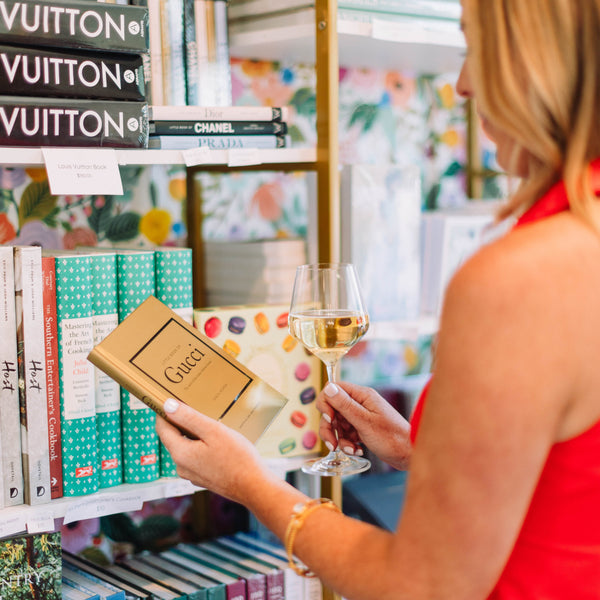 Woman in a red dress holding a glass of wine and a Gucci box in a store with shelves displaying books and luxury brands.