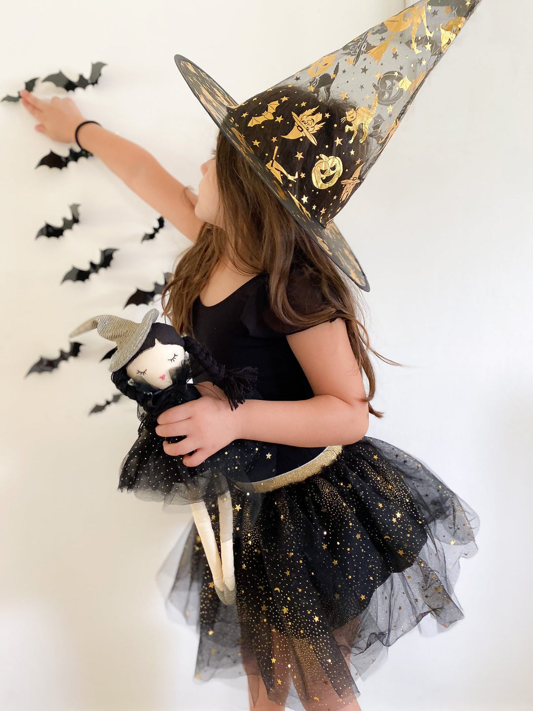 A young girl wearing a black and gold witch costume with a pointed witch hat, holding a small cat doll, posing against a white wall with hanging black bat decorations.