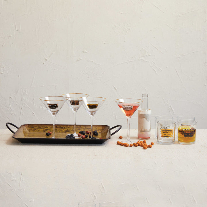 A set of four clear glass cocktail and martini glasses with gold trim and a matching gold logo, displayed on a black tray with berries, next to a pink drink, a bottle, and two small tumblers on a textured white surface.