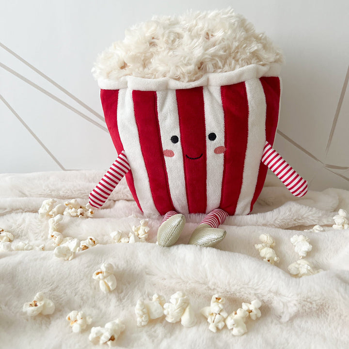 Plush toy shaped like a striped popcorn container with popped popcorn on a white surface.