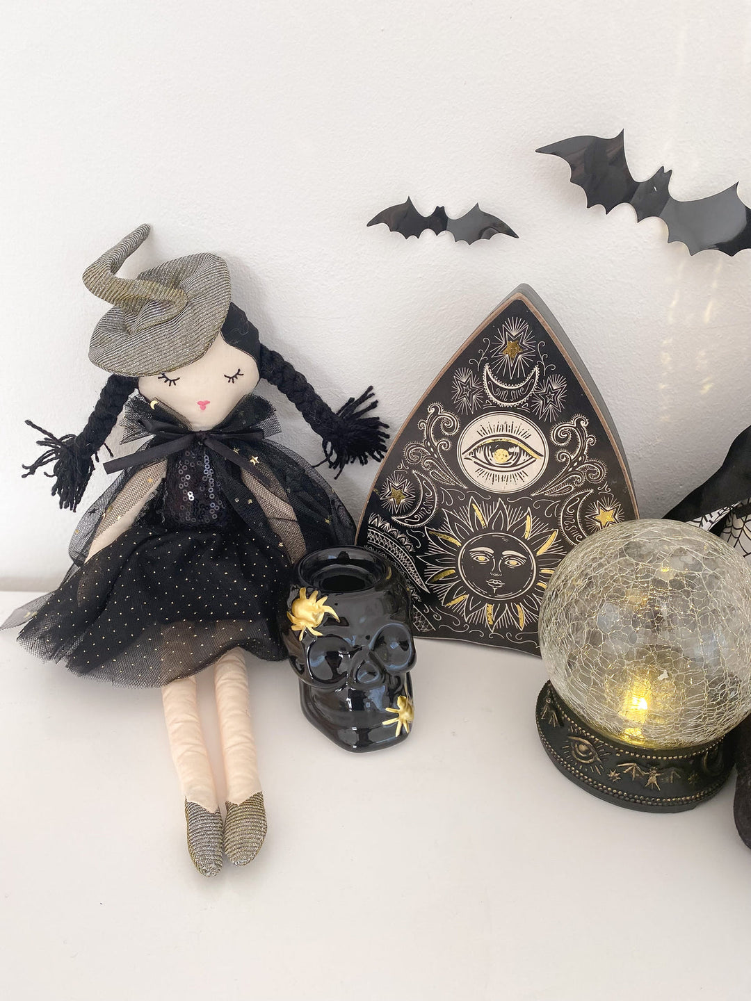 A gothic witch doll with black braids, a gray hat, and a black dress sits beside a black skull, a triangular occult-themed plaque, and a glowing spiderweb crystal ball on a white surface.