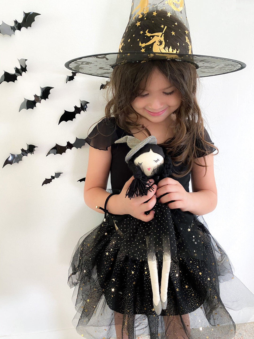 A young girl wearing a black witch costume with a black tulle hat decorated with gold stars, holding a small black-and-white doll. She is standing against a white wall with black bat decorations on the left.