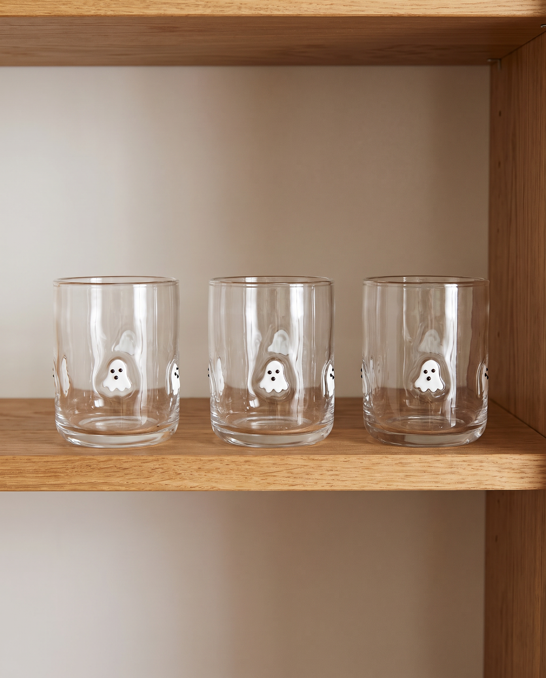 Three clear glass tumblers with white ghost illustrations, displayed on a wooden shelf against a neutral beige background.